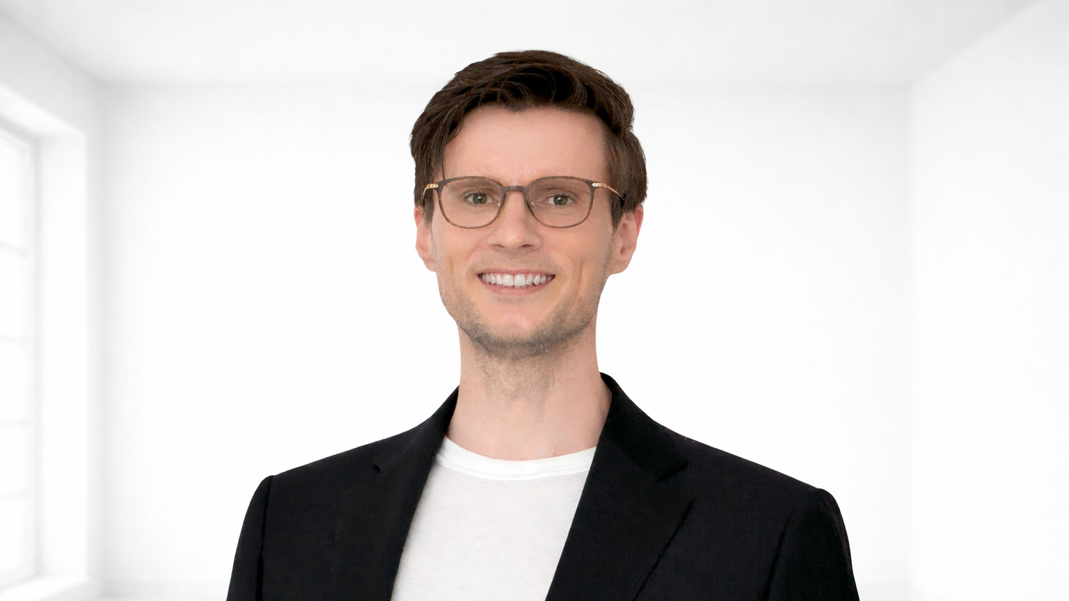 Portrait d’un homme souriant avec des lunettes, une veste noire et un t-shirt blanc, devant un fond clair et neutre.