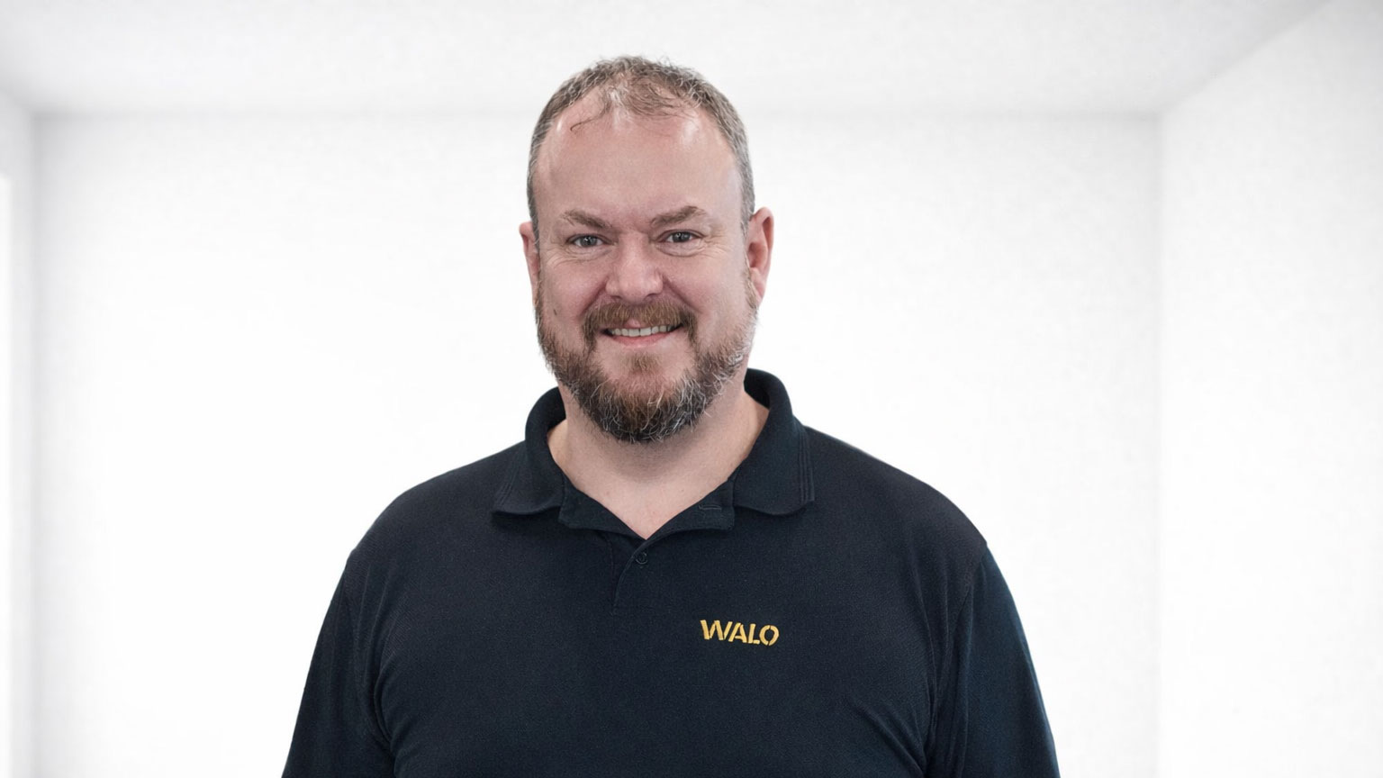 Portrait of an employee with a beard wearing a black WALO polo shirt against a light background.
